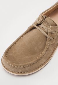 Scarpa mocassino in suede marrone con punta rotonda piatta, cuciture decorative e chiusura con lacci. Texture liscia e colore neutro.
