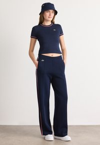 Marineblaues Crop-Top- und Hosen-Set mit roten Streifenakzenten. Verfügt über einen gerippten Halsauschnitt und ein Logo auf der Brust und den Hosen. Kombiniert mit einem passenden Bucket Hat.