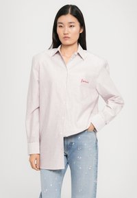 Mulher com cabelo comprido e escuro, vestindo uma camisa botão oversized, rosa e branca às riscas, e jeans azul claro com pequenas flores cor-de-rosa.