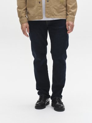 Vaqueros de mezclilla azul oscuro con un corte recto, usados con una chaqueta de lona beige y zapatos de cuero negro con superficie texturizada.