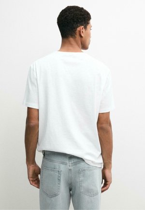 Uomo di spalle che indossa una maglietta bianca semplice a maniche corte e jeans grigio chiaro su uno sfondo bianco uniforme.