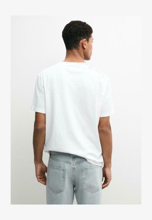 Ein Mann, der von hinten zu sehen ist, trägt ein schlichtes weißes Kurzarm-T-Shirt und hellgraue Jeans vor einem schlichten weißen Hintergrund.