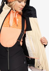 Snehová bunda s čiernym vonkajším plášťom, béžovými akcentmi a oranžovým vzorovaným vnútrom. Obsahuje zipsy a nastaviteľné popruhy.