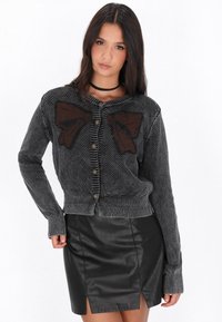 Cardigan tricotat gri cu un design în formă de fundiță maro, dotat cu nasturi aurii și manșete cu model în telecomandă. Asortat cu o fustă mini din piele neagră.