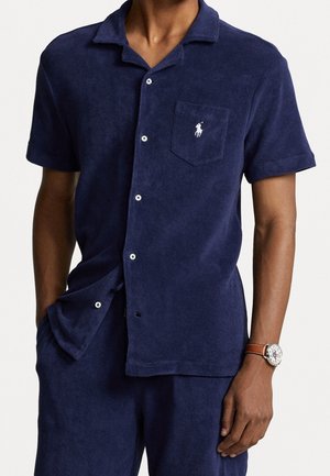 Homme portant une chemise bleu marine à manches courtes boutonnée et un pantalon, avec un petit logo blanc sur la poche poitrine et une montre-bracelet avec un bracelet marron.