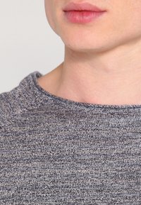 Grå stickad tröja med en texturerad yta och rund halsringning, med subtila färgvariationer och ett tätt stickat mönster.