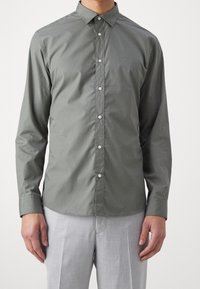 Camicia a maniche lunghe con bottoni in cotone verde oliva, dalla texture liscia, colletto classico e bottoni bianchi, abbinata a pantaloni grigi chiari.