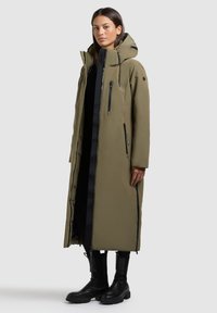 Lang olivengrøn parka med hætte, fuld lynlås, sidelommer og sorte lynlåsdetaljer. Har en polstret design og lige pasform.