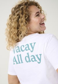 Camiseta blanca de manga corta con texto en teal que dice "Vacay All day" en la parte de atrás, con un corte relajado y suave tela de algodón.