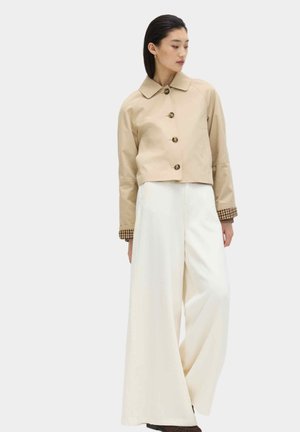 Vrouw draagt een beige jasje met knopen, geruite manchetten en brede witte broek, staand tegen een effen lichte achtergrond.