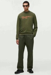 Sweatshirt verde-oliva e calças combinando feitas de tecido macio, com o logótipo "Karl Lagerfeld Jeans" em laranja. Sapatos desportivos pretos.