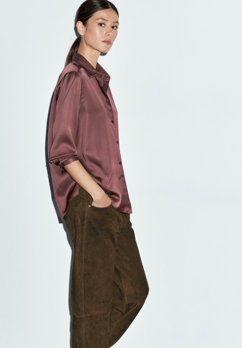 Camicia di raso bordeaux con colletto e bottoni, abbinata a pantaloni in suede marrone scuro. L'outfit presenta una vestibilità comoda e tasche laterali.