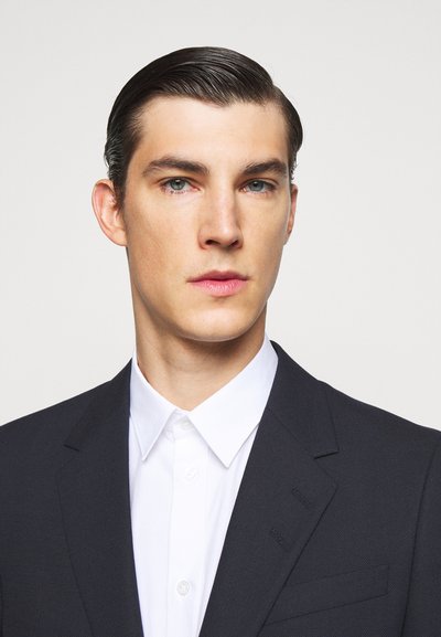 Blazer azul-marinho feito de tecido texturizado, apresentando lapelas e três botões em destaque, combinado com uma camisa branca de colarinho.