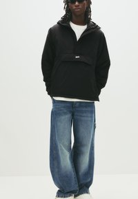 Jachetă ușoară neagră cu glugă, buzunar frontal și logo, asociată cu blugi evazați decolorati de culoare albastră și adidași albi.