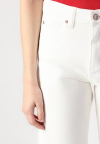 Vita denimjeans med texturerad yta, femfickorsdesign och detaljer i roséguld på knappen. Handen vilar på låret.