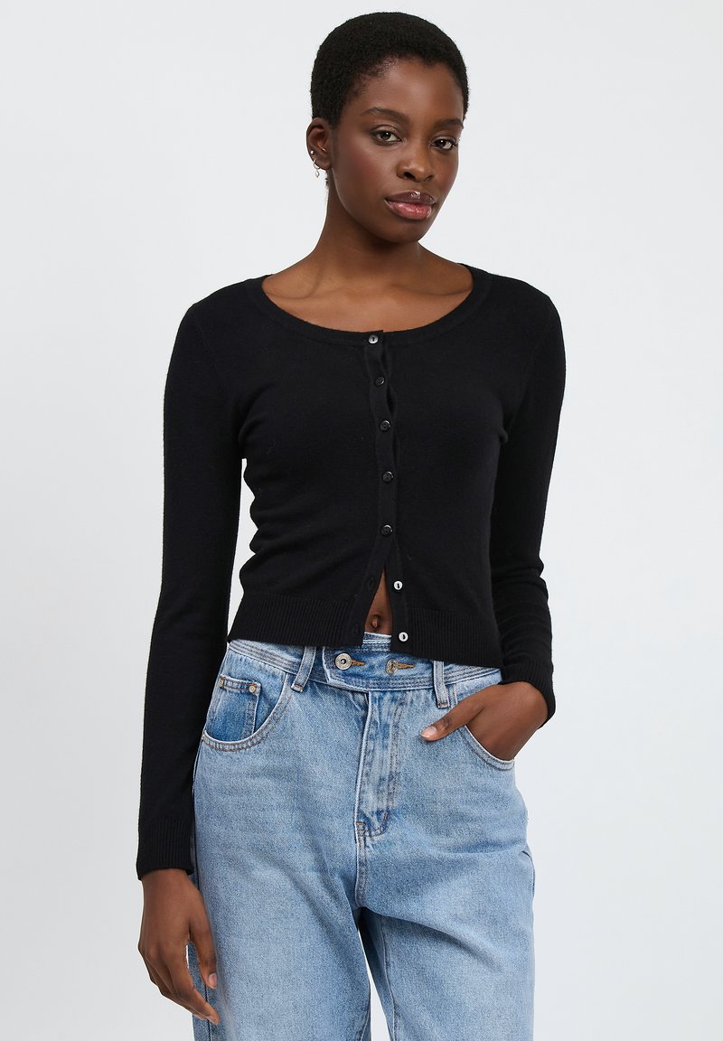 Schwarze, gekürzte Strickjacke mit langen Ärmeln, rundem Ausschnitt und einer Reihe von Knöpfen vorne, kombiniert mit hellblauen, hochsitztenden Jeans.