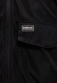 Υφασμα μαύρου μπουφάν με φερμουάρ στα αριστερά και τσέπη με καπάκι στα δεξιά που εμφανίζει μια ετικέτα "Barbour International".