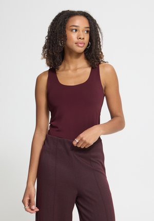 Jeune femme aux cheveux bouclés portant un haut bordeaux sans manches et un pantalon taille haute assorti, regardant sur le côté sur un fond uni.