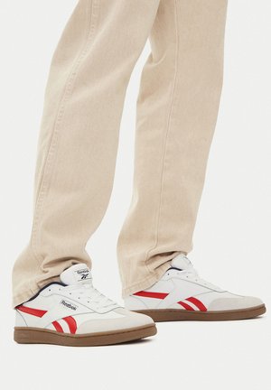 Białe sneakersy Reebok z czerwonymi akcentami, z teksturowaną cholewką i gumową podeszwą. W zestawie z jasnobeżowymi, zwężanymi spodniami.