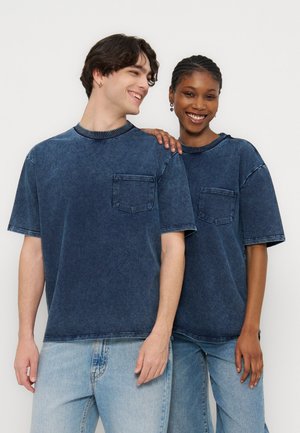 Du žmonės šypsosi, vilki tamsiai mėlynas, kišenėmis išsiuvines marškinėlius ir šviesiai mėlynas džinsus, vienas laiko ranką ant kito peties.