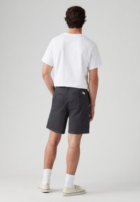 Pantaloni scurți gri dintr-un material cu o textură netedă, având două buzunare la spate cu fermoar, purtați cu un tricou alb și încălțăminte deschisă.