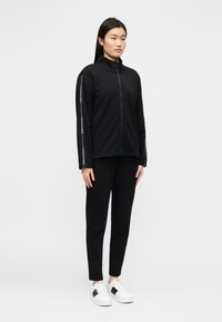 Zwarte jas met rits en hoge kraag en strepen aan de zijkant, gecombineerd met taps toelopende zwarte broek en witte sneakers met zwarte accenten.