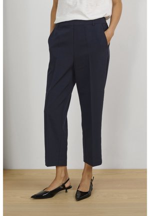 Kobieta ubrana w granatowe, skrócone, dopasowane spodnie z przednimi zakładkami, białą bluzkę i czarne buty slingback na niskim obcasie, stojąca na drewnianej podłodze.