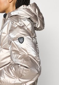 Metāliski bēša pūkaina jaka ar kapuci, ar ielocītu tekstūru, melnu EA7 logotipa uzšuvumu uz piedurknes un rāvējslēdzēja aizdari.