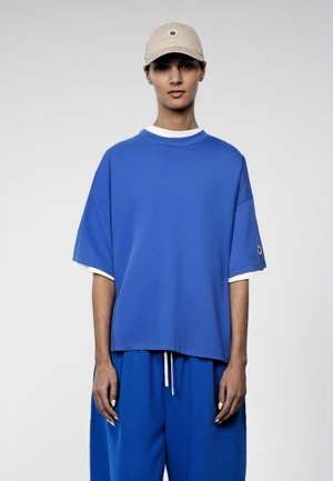 Femme debout face à l'avant, portant une casquette beige, une chemise ample à manches courtes bleue sur un haut blanc, et un pantalon bleu assorti avec un cordon de serrage.