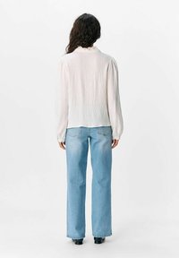 Blusa bianca a maniche lunghe, texturizzata, con colletto arricciato, abbinata a jeans alta vita azzurri e scarpe nere.