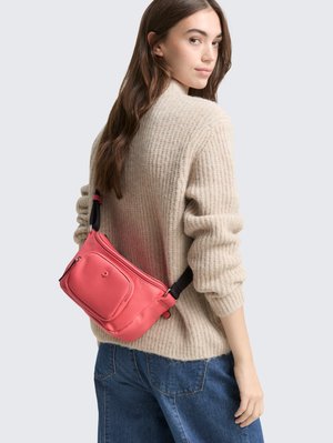 Jauna moteris, apsirengusi smėlio spalvos megztiniu ir mėlynomis džinsomis, nešioja koralų rožinės spalvos per petį užsegamą rankinę, žiūrėdama per petį.