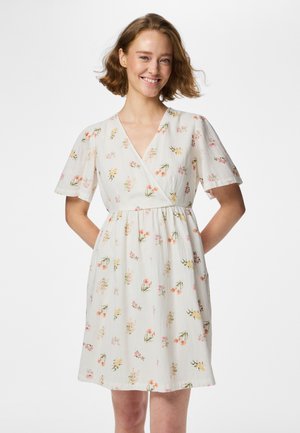 Lachende vrouw met kort golvend haar, gekleed in een witte jurk met een geel-roze bloemenpatroon, handen achter haar rug.