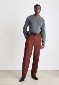 Grå långärmad skjorta, löst sittande vinröda byxor, svart bälte med metalldetaljer och svarta läderskor, står på en neutral bakgrund.