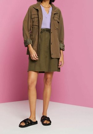 Femme portant une veste olive et une jupe, haut lilas, sandales noires, debout contre un mur rose.