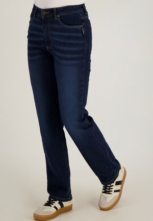 Persoon die donkerblauwe jeans met rechte pijpen en witte sneakers met zwarte strepen draagt, staand met één been licht gebogen.