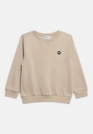Beige long-sleeve crewneck sweatshirt with ribbed cuffs and hem, small black circular logo on the left chest.