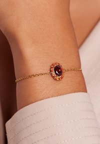Gouden armband met een centrale ovale rode edelsteen omringd door kleinere oranje en amberkleurige stenen, aan een verfijnde ketting. Textuur van de huid zichtbaar.