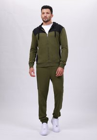 Olivgrüner Hoodie und Jogger-Set aus weichem Material, mit schwarzem oberen Einsatz, Reißverschluss und weißen Sneakern. Lässige Passform.