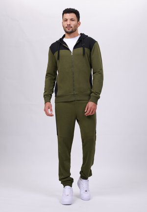 Olijfgroene hoodie en jogger set van een zachte stof, met een zwart bovenpaneel, ritsluiting en witte sneakers. Relaxte pasvorm.