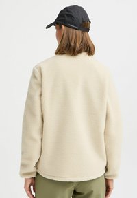 Veste en polaire beige avec une surface texturée, col rond et épaules légèrement tombantes, accompagnée d'une casquette noire en arrière-plan.