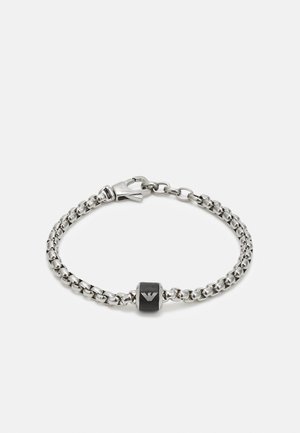 Sølvkjedearmbånd med et svart rektangulært anheng med logo, festet med en lås. Lenkene er glatte og metalliske.