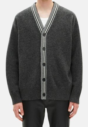 Homme portant un cardigan gris foncé boutonné avec un liseré à carreaux noir et blanc par-dessus une chemise blanche et un pantalon noir.