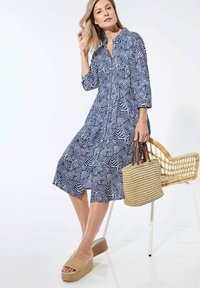 Blau-weiß gemustertes Hemdkleid mit langen Ärmeln, knielang, mit vorderer Knopfleiste und seitlichen Schlitzen. Gewebte Handtasche und Plateausandalen.