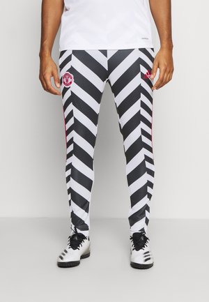 Hombre vestido con pantalones de entrenamiento de Manchester United con un patrón en zigzag en blanco y negro y botas Adidas blancas, de pie sobre un fondo liso.