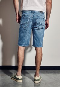 Pantalones cortos de mezclilla azul claro con un corte recto, bolsillos traseros y una cinturilla texturizada. Combinados con zapatos verdes, de pie frente a un fondo neutro.