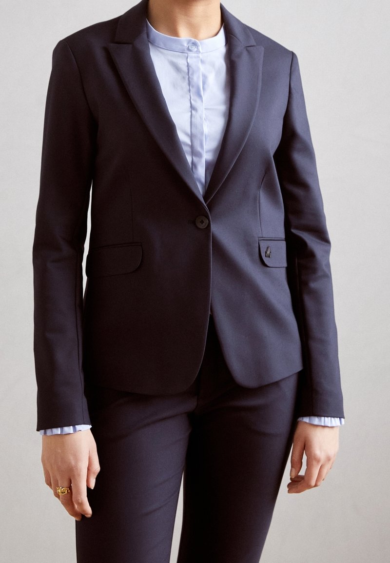 Blazer bleu marine à coupe ajustée, à un bouton, poches latérales et un emblème subtil. En dessous, une chemise bleu clair à col avec des poignets à volants.