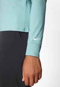 Light blue athletic long-sleeve top with ribbed cuffs; displays a white Nike logo on the left sleeve. Worn with black fitted bottoms.