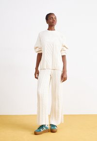 Jersey de punto crema con diseño de trenzas y mangas abullonadas, combinado con pantalones de pierna ancha a juego, luciendo con zapatillas en color azul verdoso.