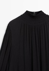 Haut noir à manches longues avec un col montant froncé. Tissu texturé avec un détail de plissage, doté d'une fermeture éclair invisible au dos.