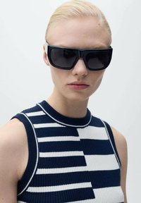 Occhiali da sole neri con montatura larga; indossati con un top senza maniche in maglia con motivi a righe blu navy e bianchi.